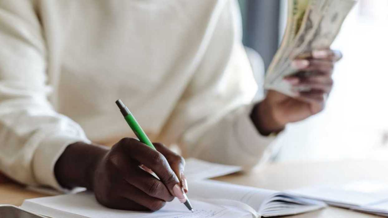 Cropped shot of a woman holding money while sitting at the table and writting expenses.