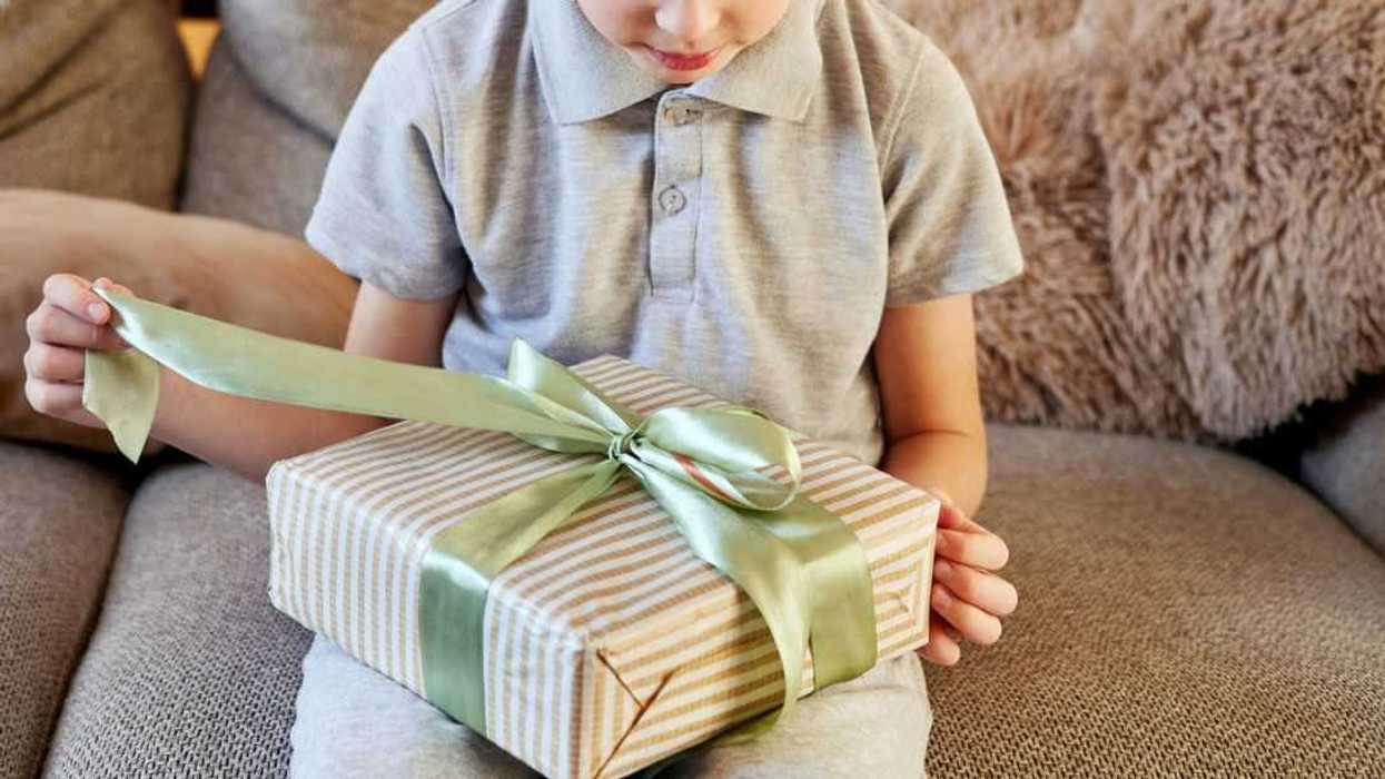 Curious young boy on a comfy sofa carefully unties a light green ribbon on a striped gift box, eagerly anticipating the surprise inside during a cozy home moment.