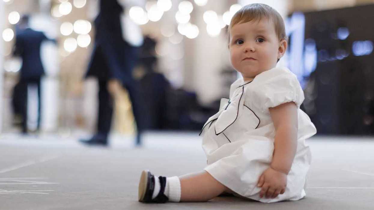 Cute baby in a white fancy dress sitting on the floor of an the event hall. Nice baby girl wearing elegant white and black dress and attractive shoes. Guests invited for wedding and waiters in the background.