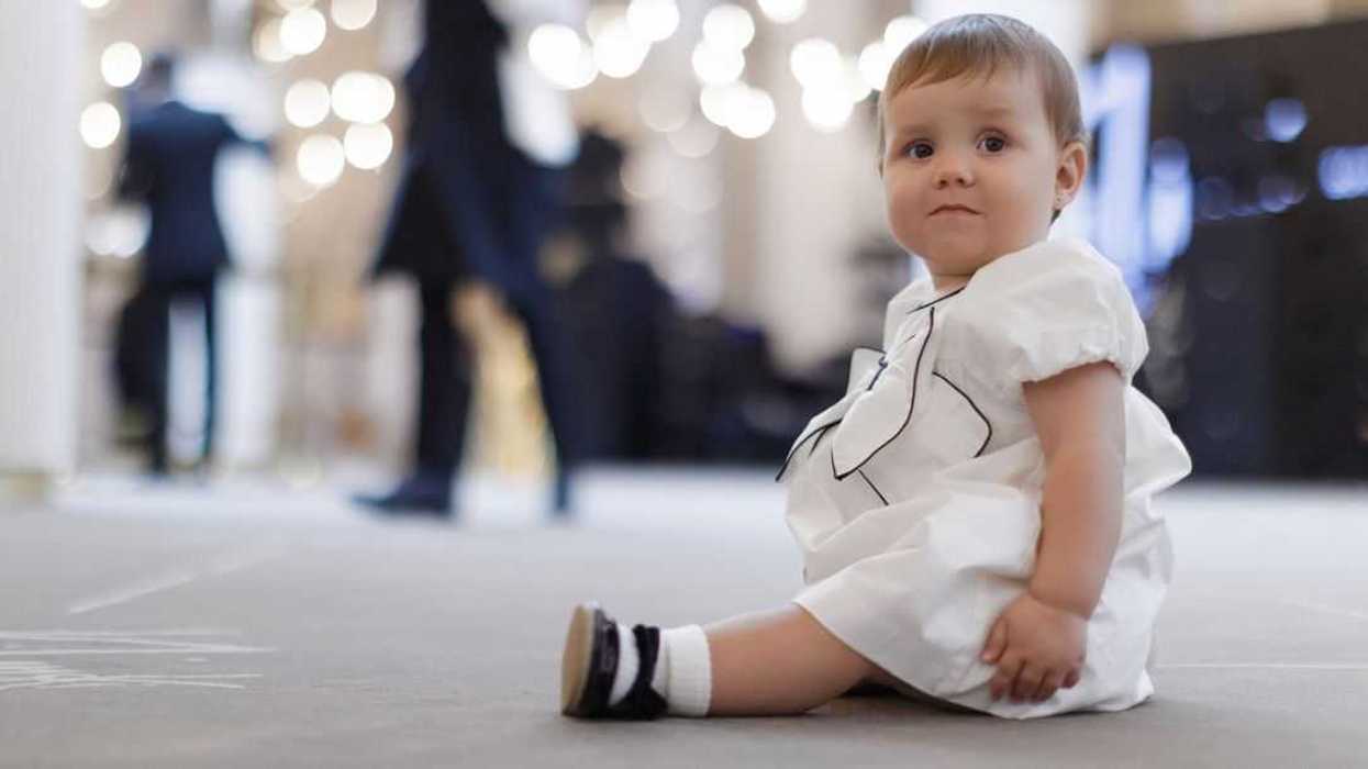 Cute baby in white fancy dress sitting at the event hall. Nice baby girl wearing elegant white and black dress and attractive shoes. Guests invited for wedding and waiters in the background.