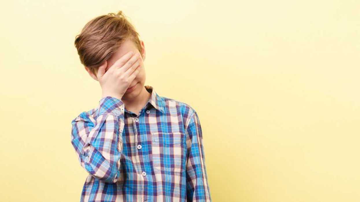 Facepalm, desperate boy standing in front of a yellow background, covers his face with his hand.