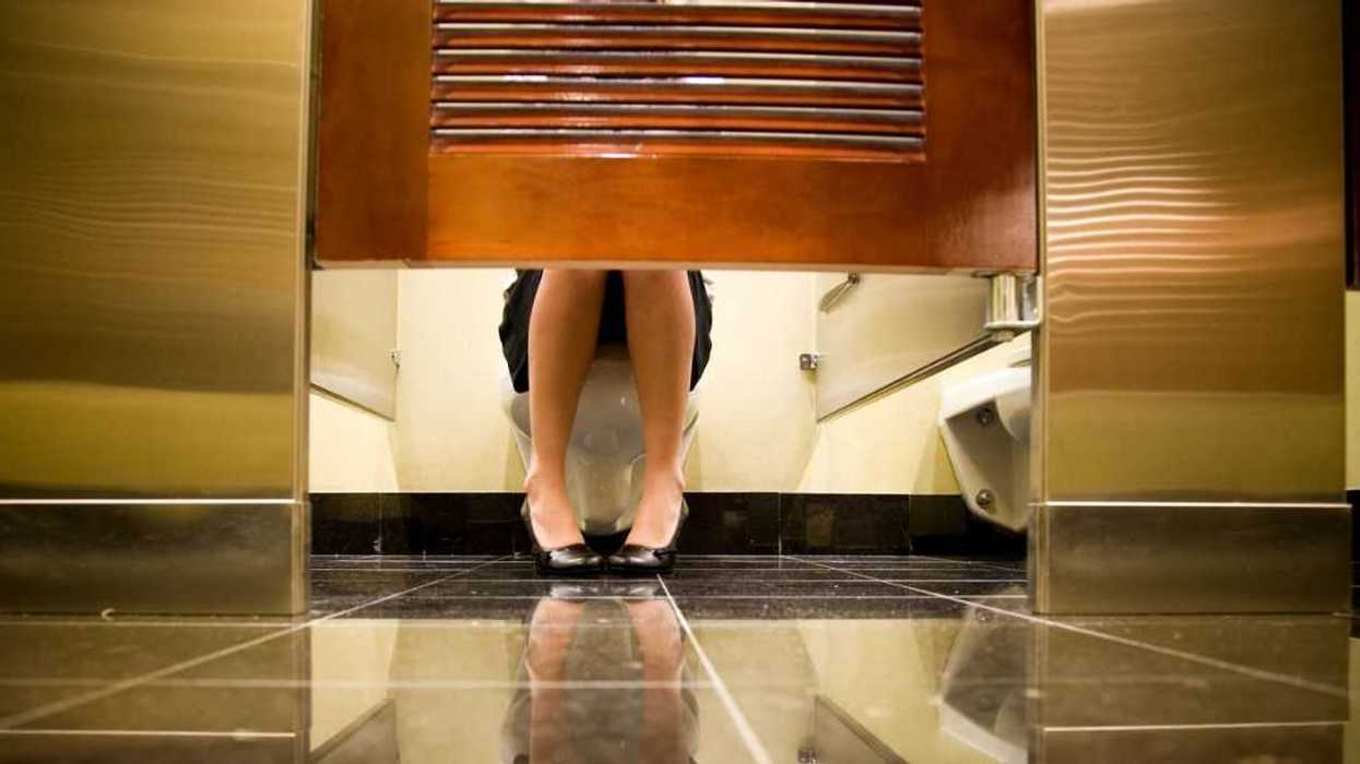 Female woman shown sitting on a toilet seat in a public washroom. Reflection of female is shown on the floor. Woman wearing business attire and wearing high heels. Feet are pointing towards each other.