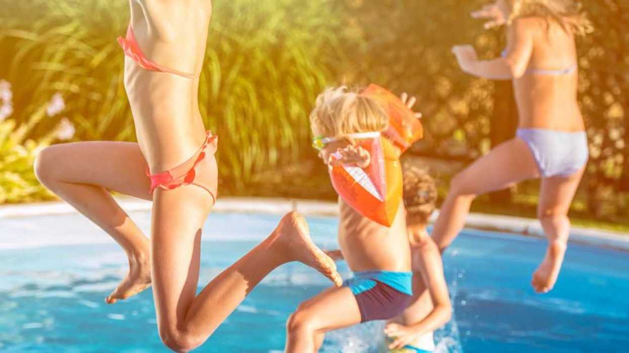 Four kids, one with water wings jumping into a pool in the backyard.