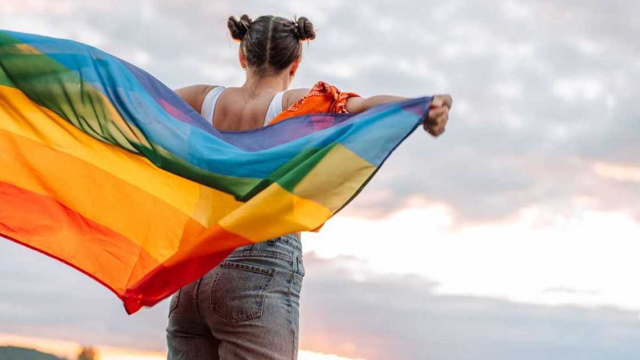 Girl standing back and holding rainbow flag on sky background.