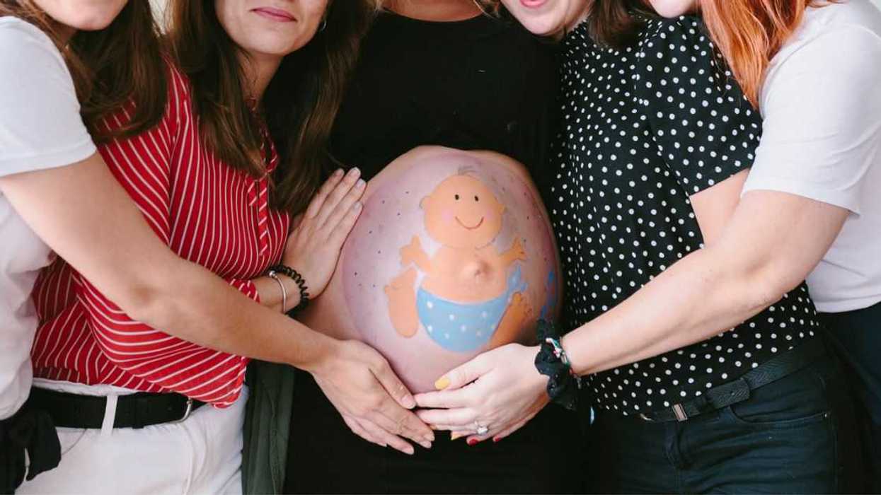 Group of smiling, female friends surrounding a pregnant woman at a baby shower. There is a baby drawn on the mother's belly.