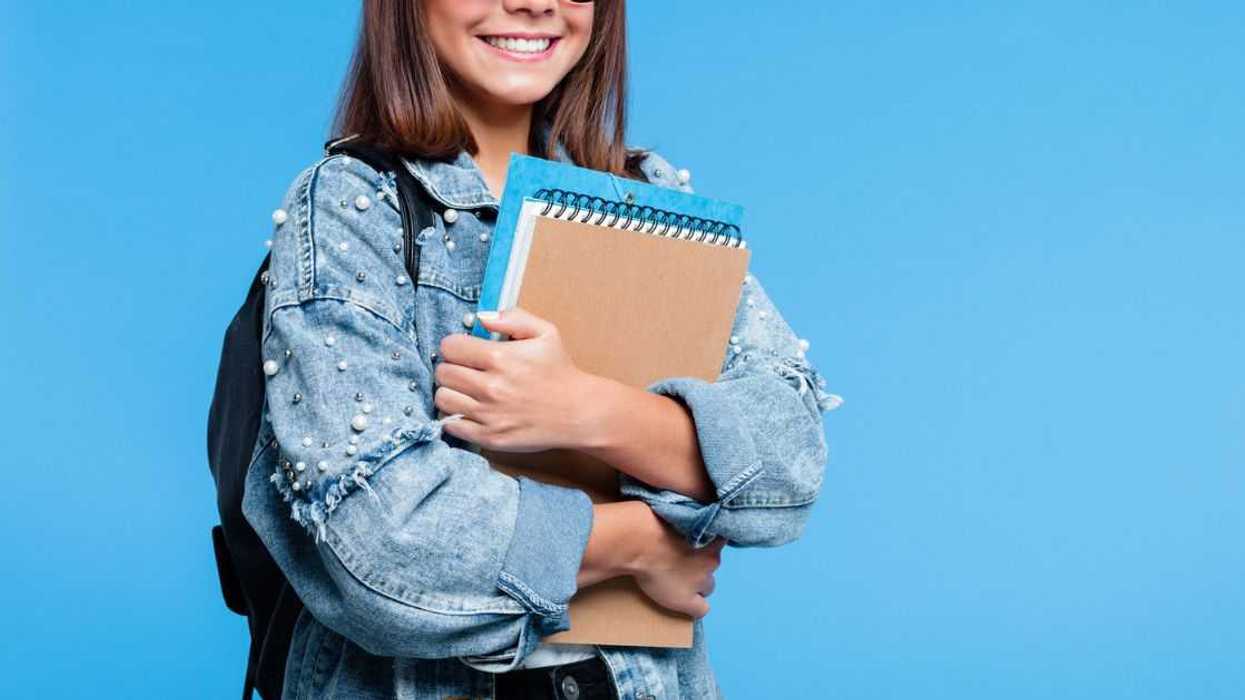 Happy cute teenage girl wearing oversized denim jacket, black eyeglasses and backpack holding books. Smiling teenager standing against blue background.