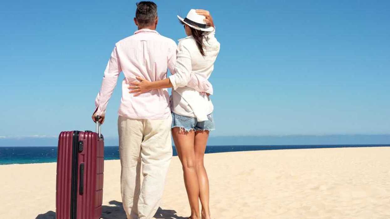 Happy traveling couple standing with suitcases on a sandy beach on a sunny summer day.