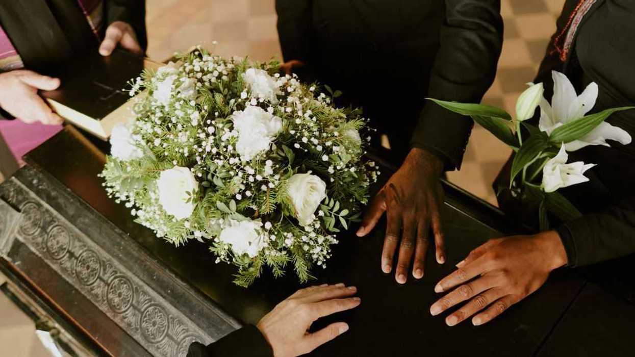 High angle shot of unrecognizable hands on black wooden coffin with white roses on top, pastor holding holy book.