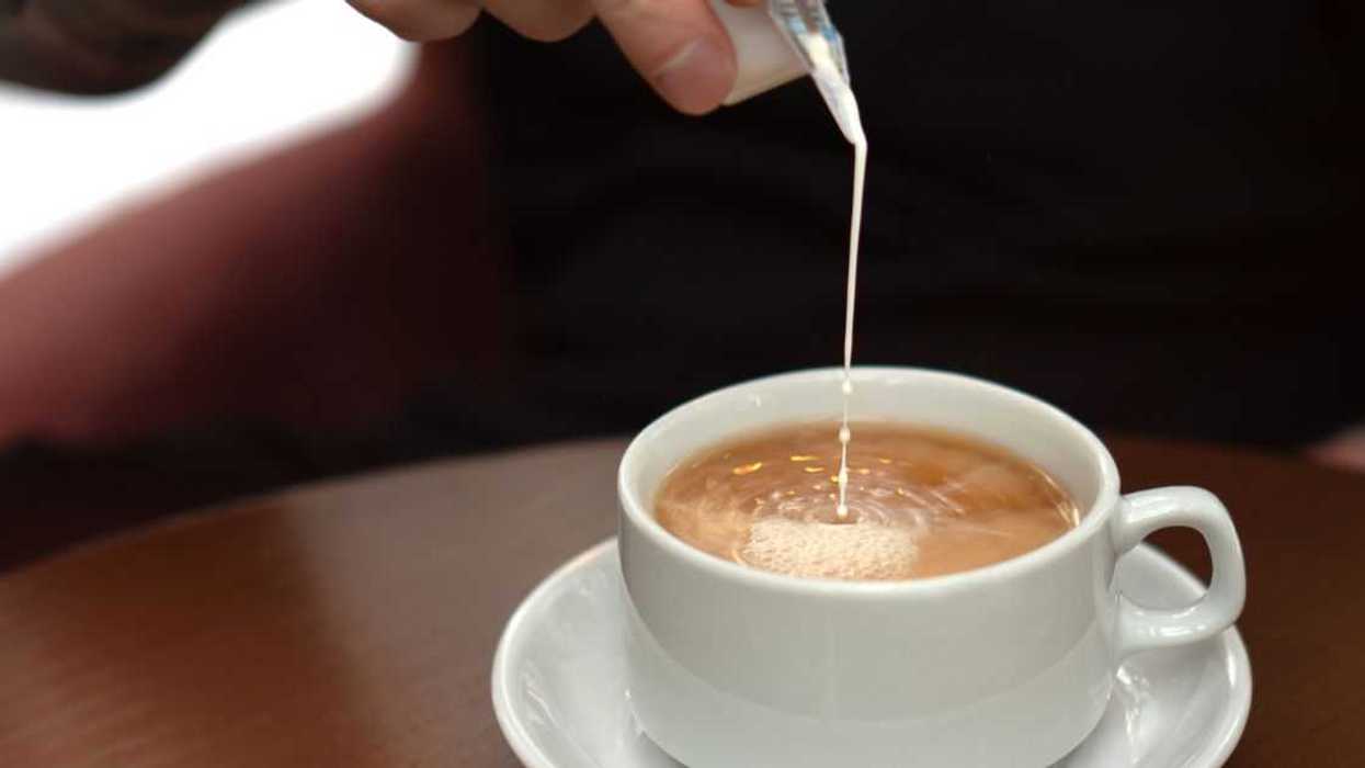 In the cozy teahouse. Male hand pouring milk into coffee in a white ceramic cup on saucer. Closeup of natural hot beverage. Front view, high angle.
