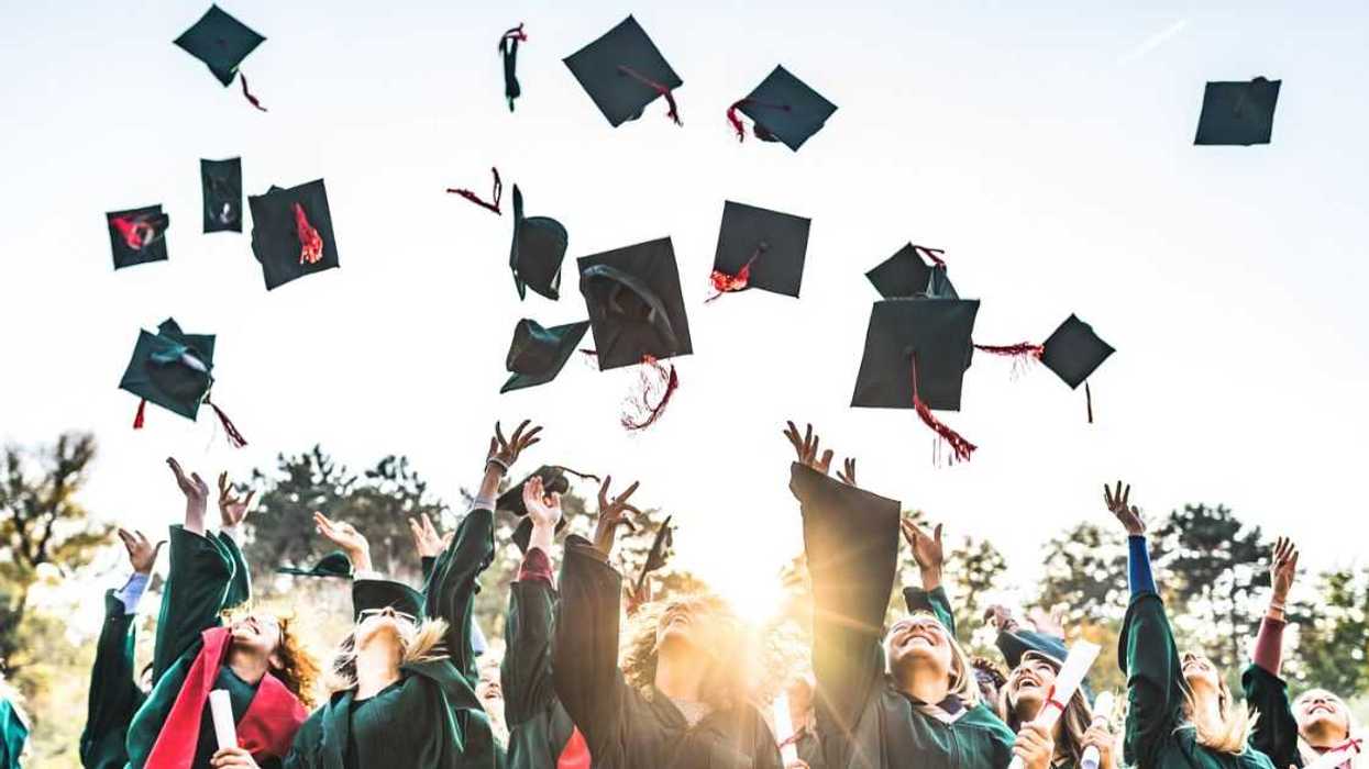 Large group of happy college students celebrating their graduation day outdoors while throwing their green caps up in the air.