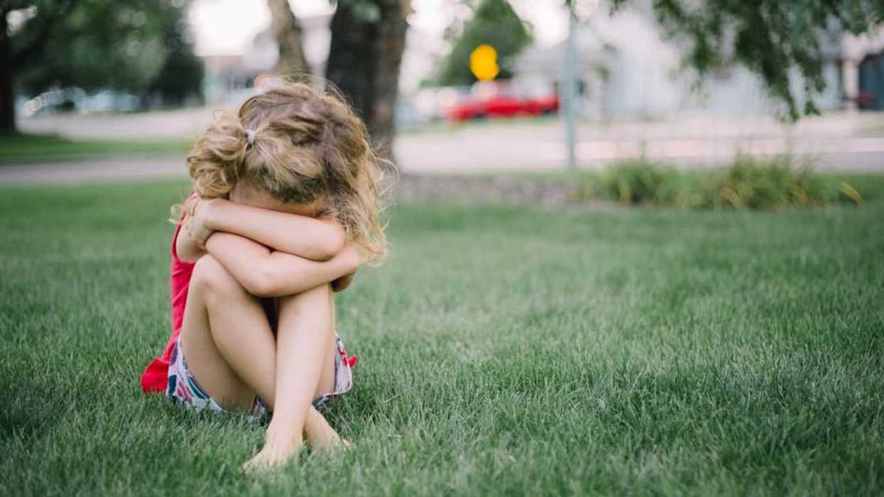Little girl with curly hair sits on grass and cries into her arms.
