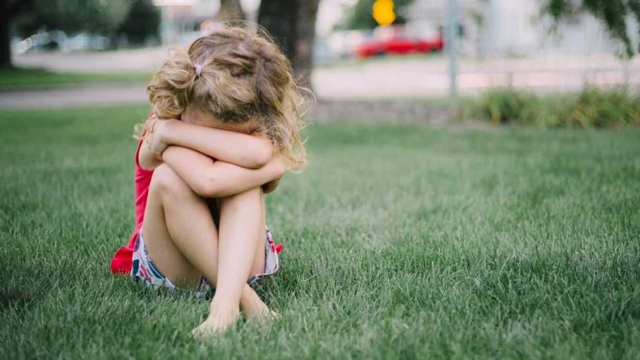 Little girl with curly hair sits on grass and cries into her arms..