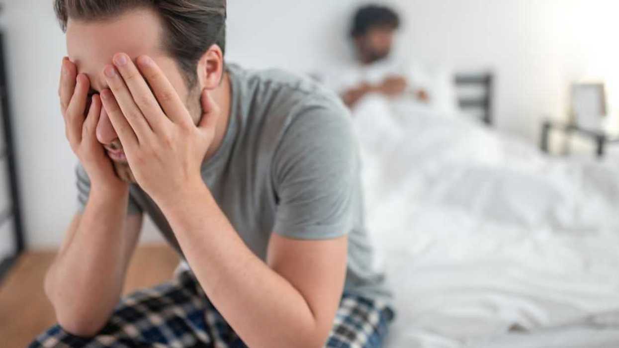 Male sitting with his eyes closed on the edge of the bed far away from his partner lying in bed.