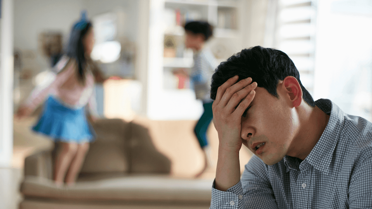 man with head in hand while children jump on furniture in background
