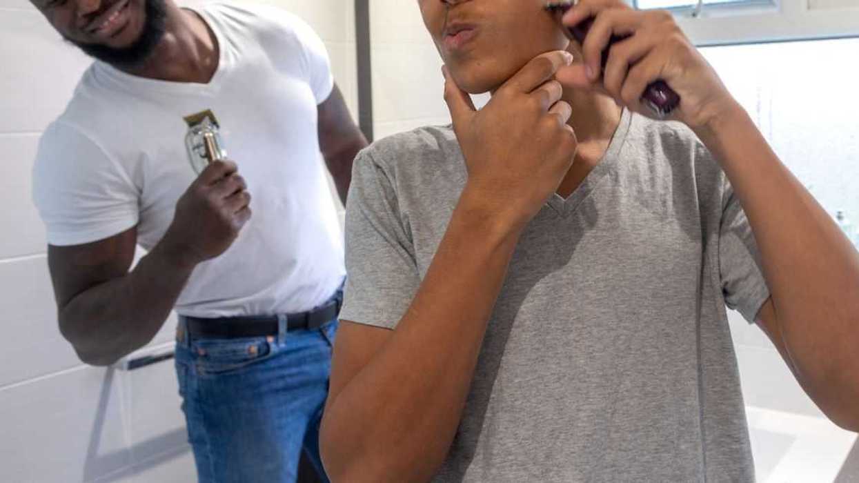 Medium point-of-view shot of a teenage boy and his father standing in the bathroom of their home in Sunderland, North East England. The boy is using an electric razor to trim his facial hair, he is distorting his face as he shaves one cheek. His father is smiling at him in the background.