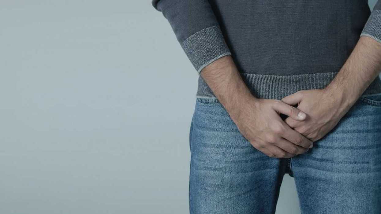 Mid-section shot of a young man holding his crotch. He's wearing light blue jeans, a gray sweater and stands against a gray background.