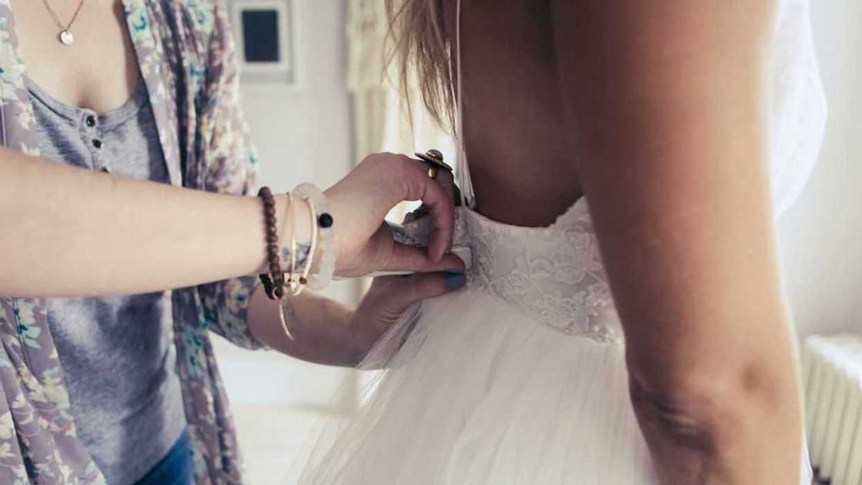 Midsection shot of a bride to be standing in an elegant wedding dress and looking at herself in the mirror during a fitting in a bridal shop, with the stylist making small adjustments to the dress.