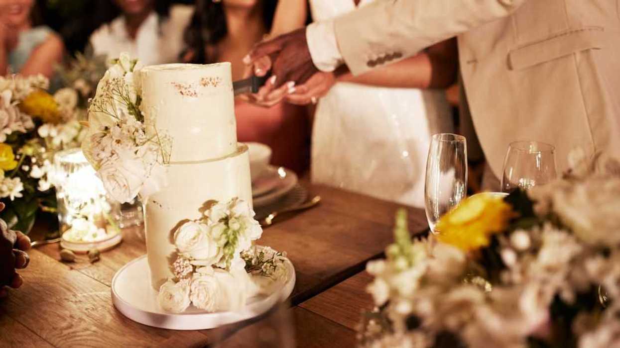 Midsection shot of a young newlywed couple cutting their wedding cake by guests at an illuminated dining table during wedding reception.