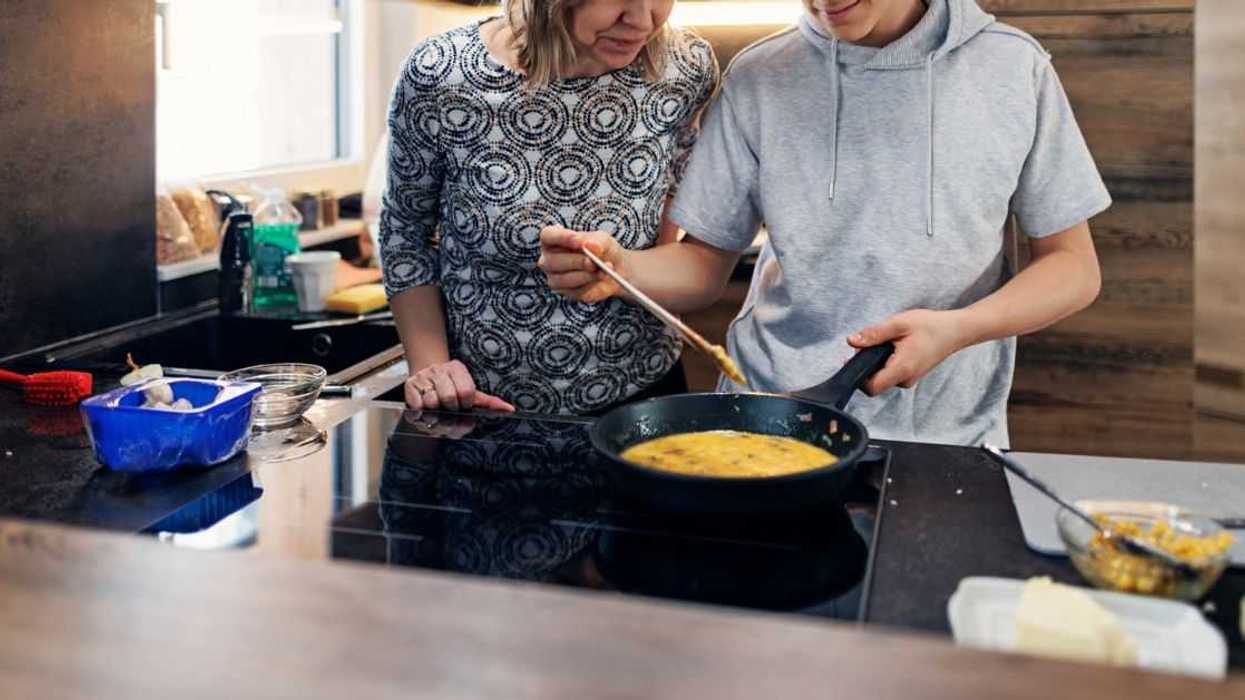 Mother and teenage son are preparing breakfast together. They are making the scrambled eggs. The boy is mixing the eggs in the frying pan.