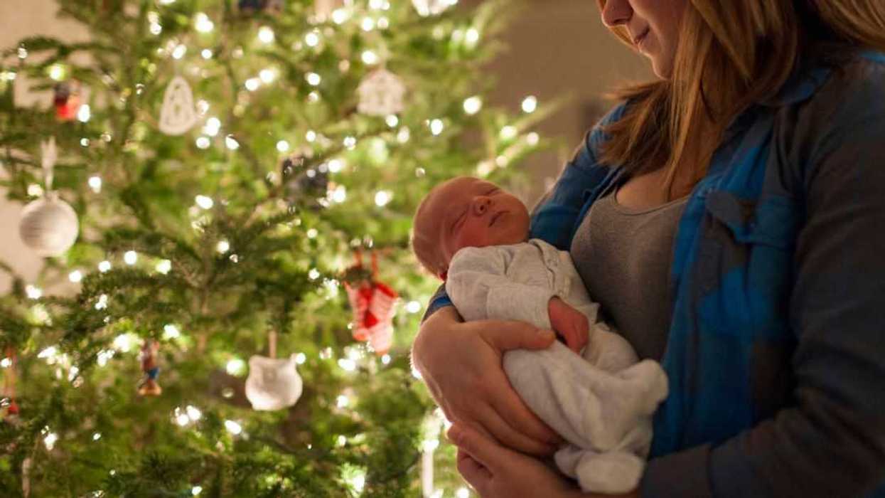 Mother carrying newborn baby while standing in front of a Christmas tree at home.