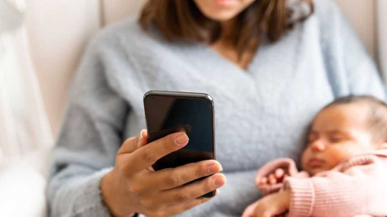 Mother using her cell phone while sitting on the living room sofa while her newborn sleeps in her arms.