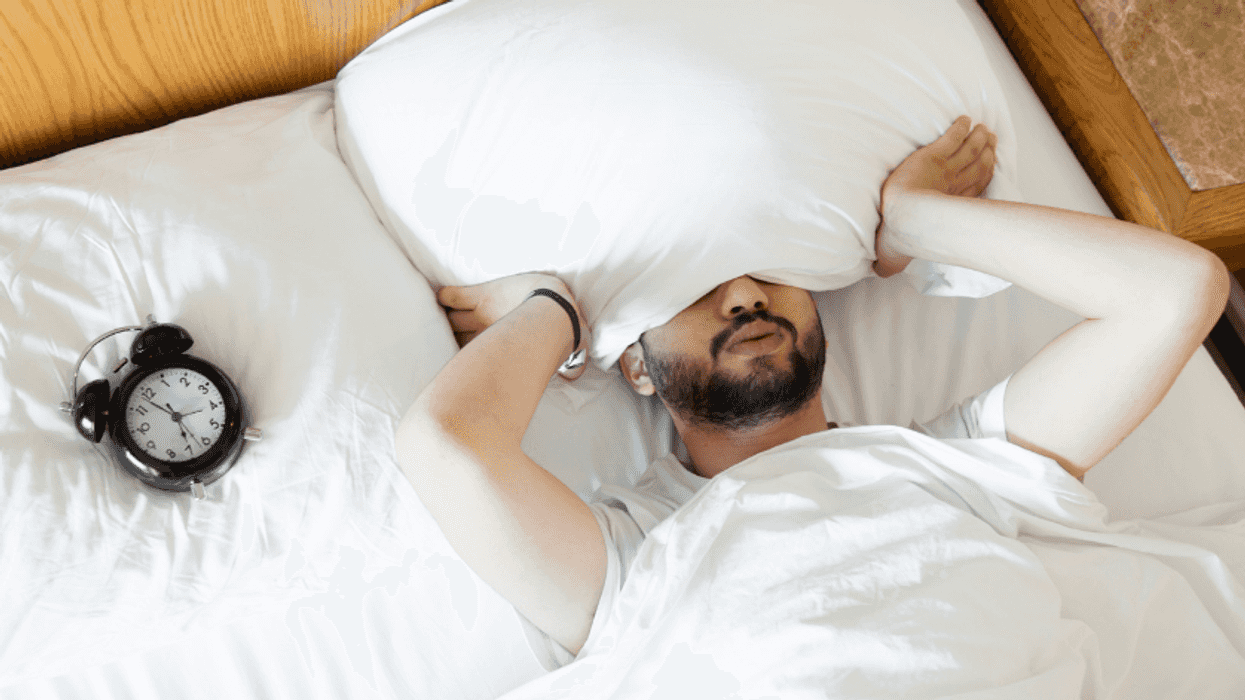person laying on bed with head under pillow with alarm clock next to them