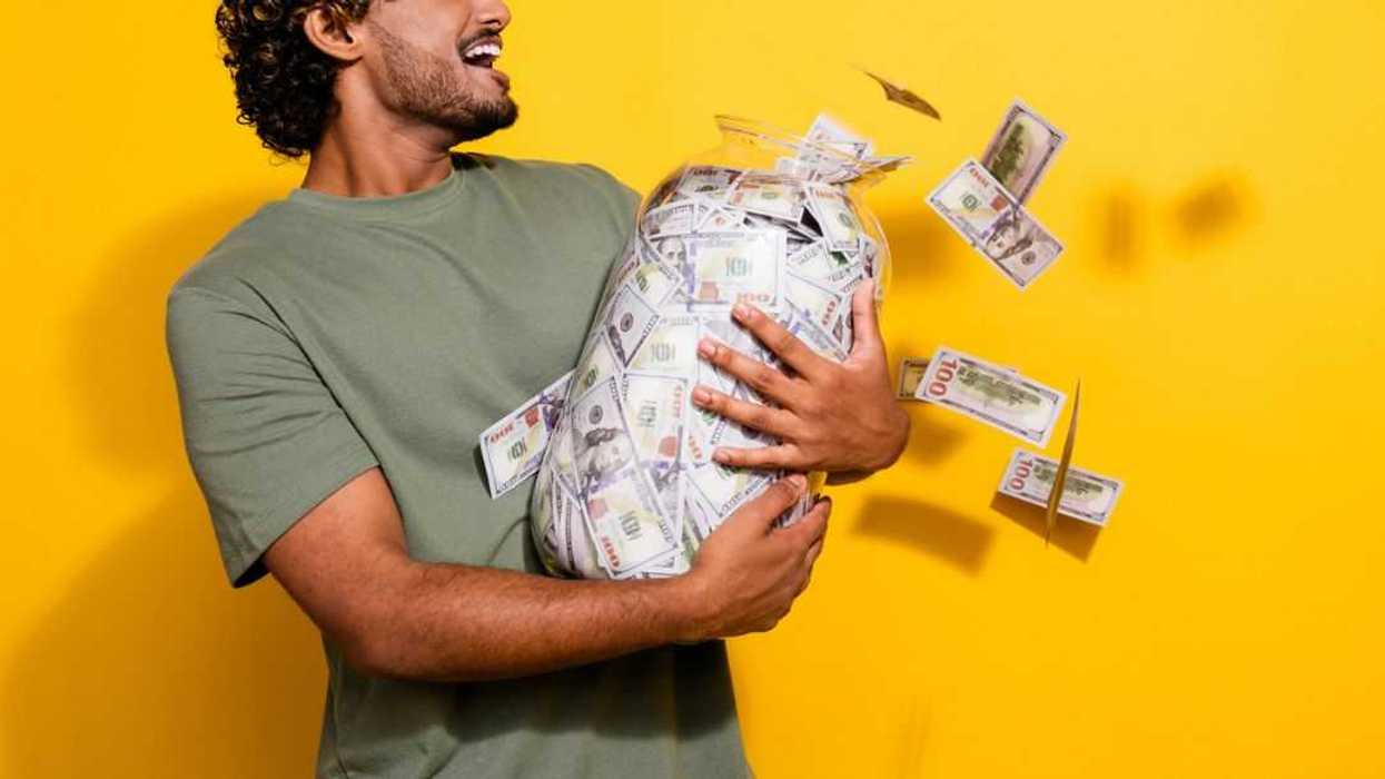Photo of an impressed guy with wavy hair, dressed in a green khaki t-shirt, holding a jar with money, looks at flying dollars isolated on yellow color background.