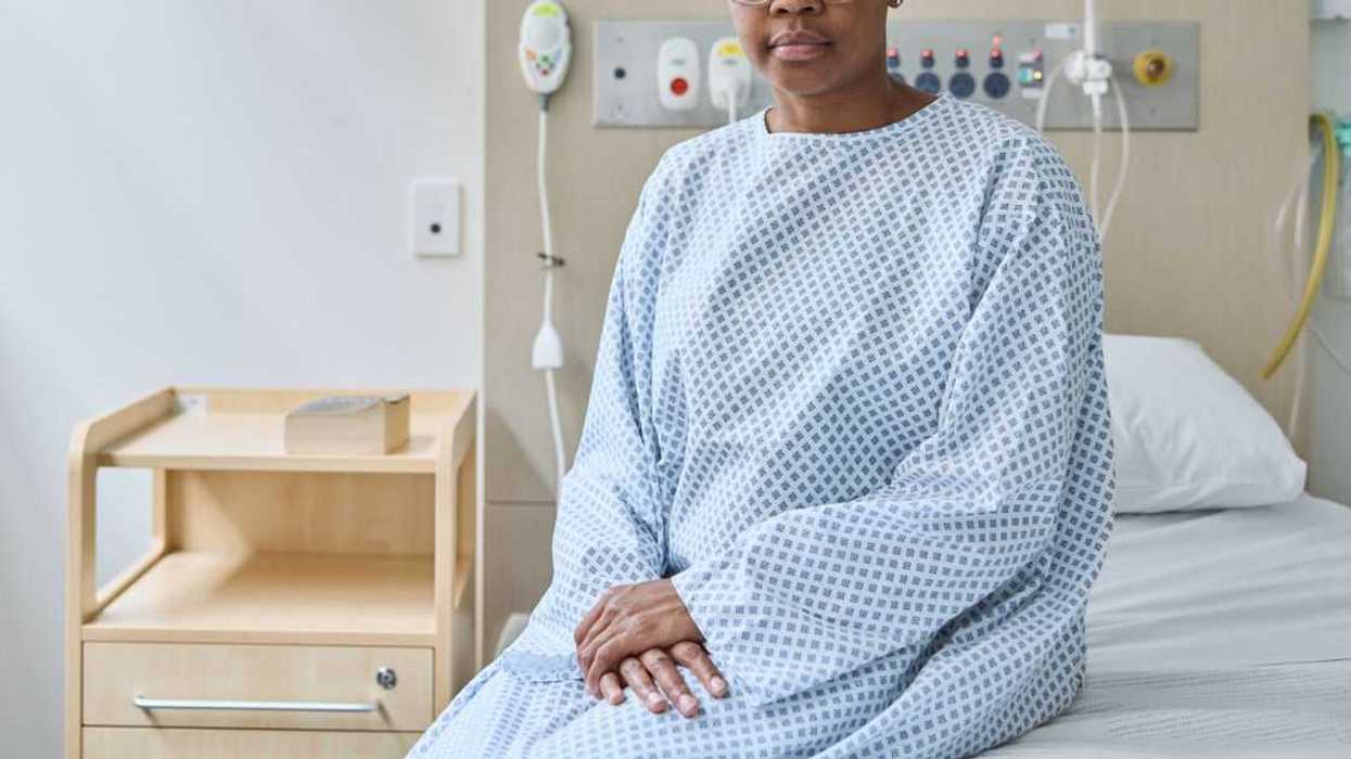Portrait of a bald woman suffering from cancer sitting on bed at hospital. Female patient is wearing medical gown. She seems to be serious.