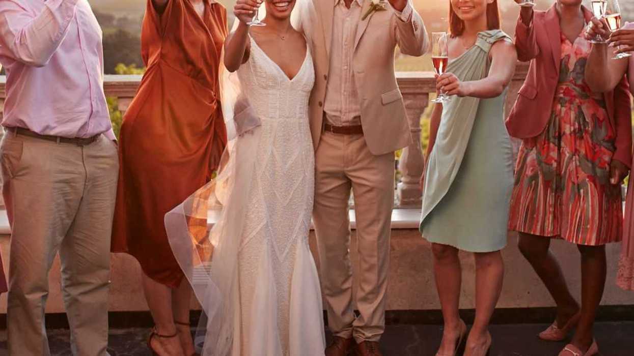 Portrait of a happy just married couple and guests toasting drinks while standing against sky at sunset.