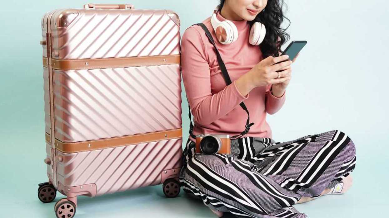 Portrait of a young, pretty girl, sitting next to a large, aluminium suitcase. She is texting and wears large headphones around her neck.