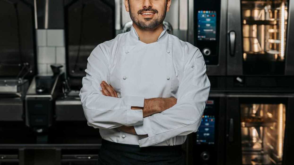 Portrait of confident, young male chef wearing a white uniform and standing with his arms crossed in the kitchen of a restaurant.