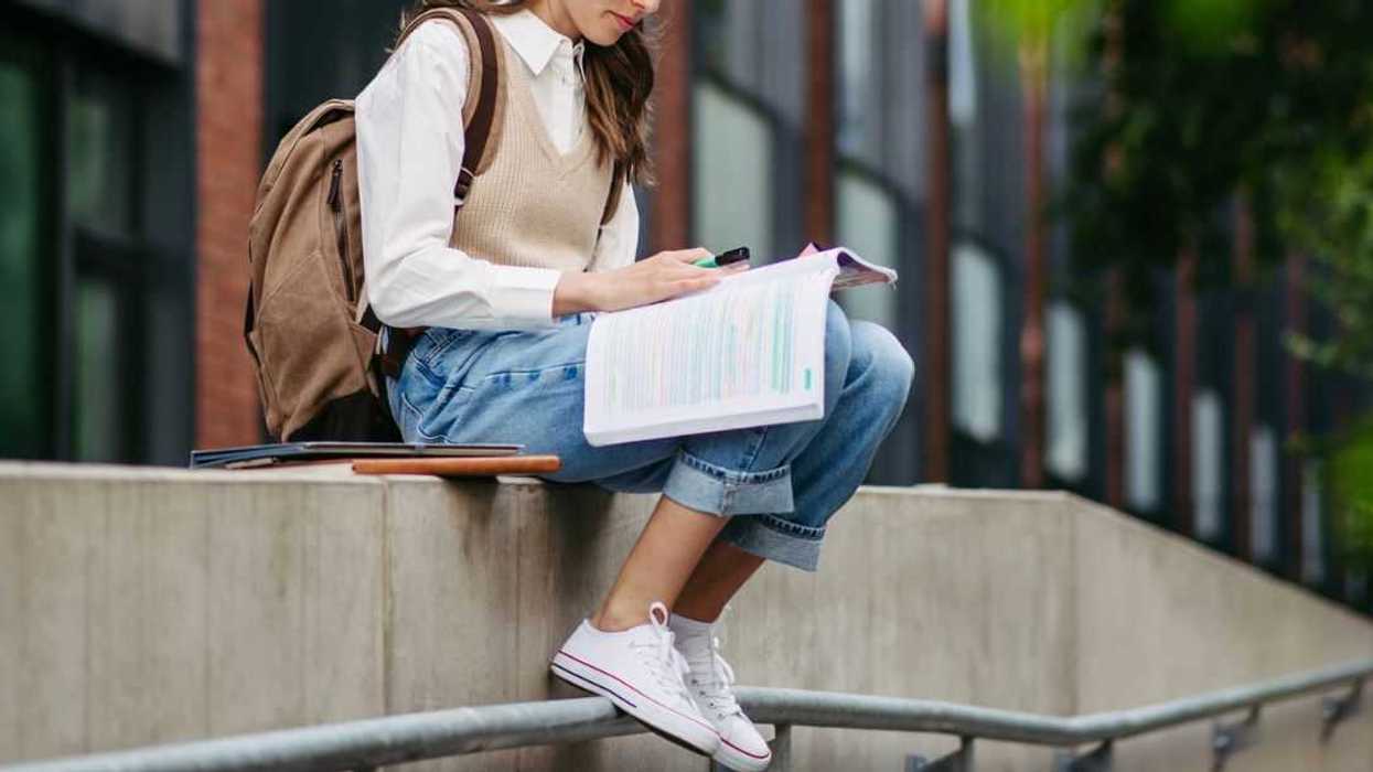 Portrait of female student sitting on low wall in front of university building, studying, reading textbook, preparing for final exam.