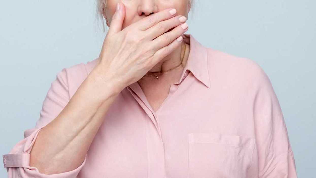 Portrait of terrified senior woman with gray hair wearing pink blouse covering mouth with hand. Studio shot, grey background.
