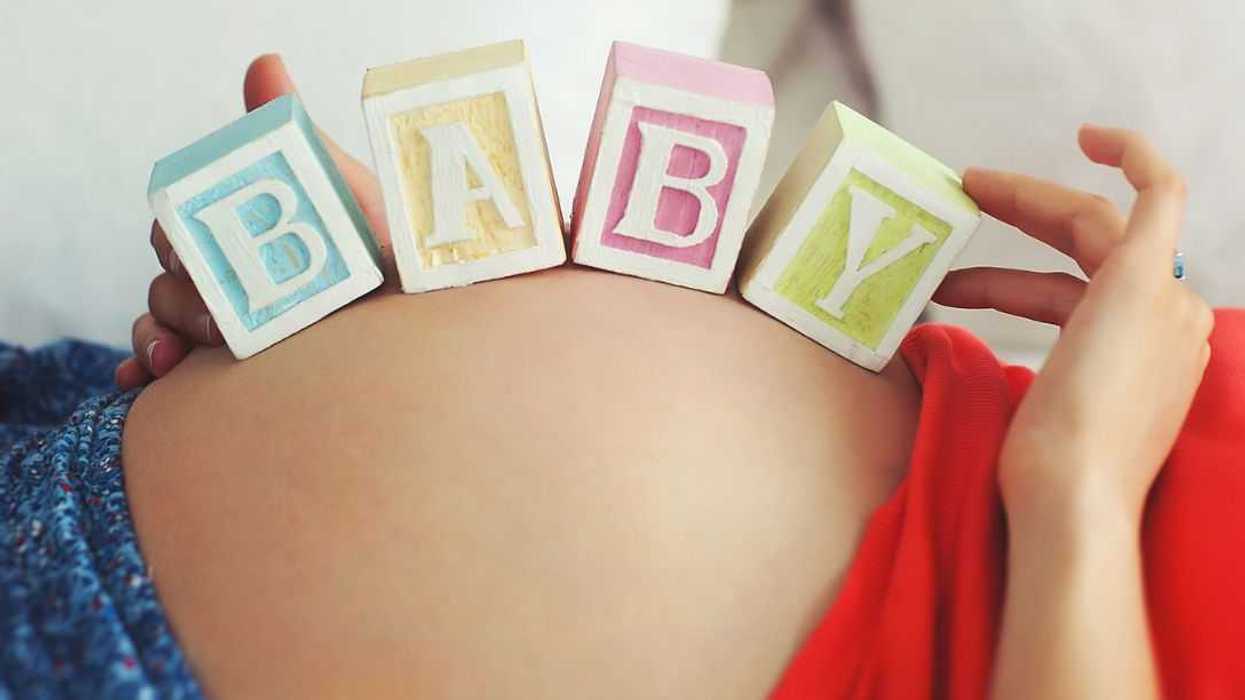 Pregnant woman balancing, with hands, wooden toy blocks with letters spelling baby on her baby bump in bed.