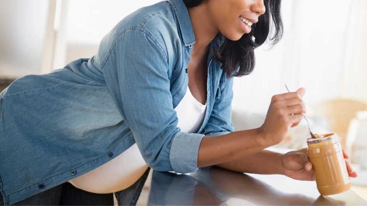 Pregnant woman leaning over a counter while eating peanut butter in the kitchen.