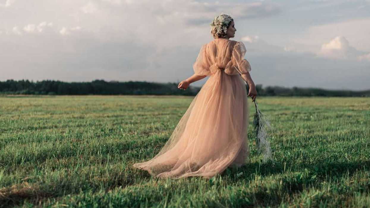 Rear view of a woman wearing vintage pink dress while walking in a field against the sky.