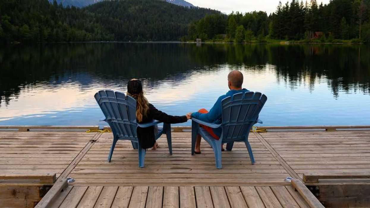 Rear view of couple relaxing on Adirondack chairs at lake. Male and female are spending leisure time together. They are on pier.