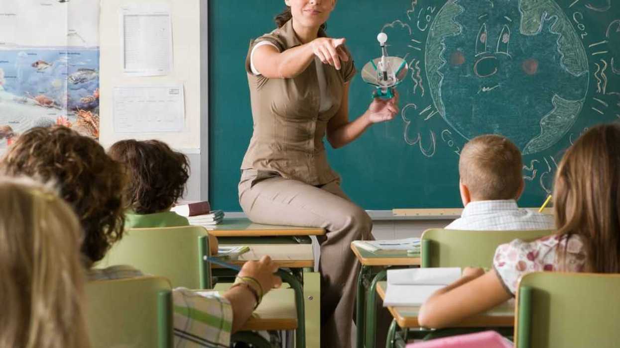 Science teacher sits on the edge of her desk, she is calling on a student and holds a light bulb.