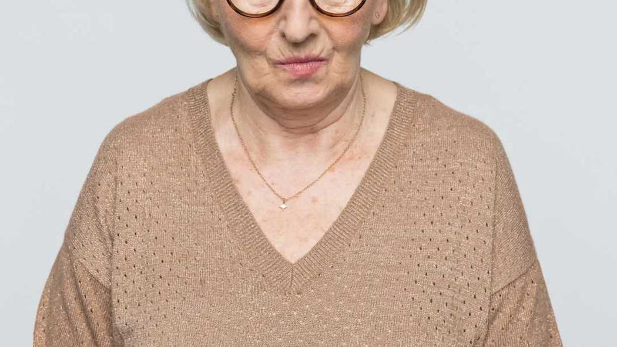Serious elderly woman with short blonde hair and round eyeglasses looking directly at camera, wearing beige V-neck sweater and delicate necklace, isolated on light gray background.