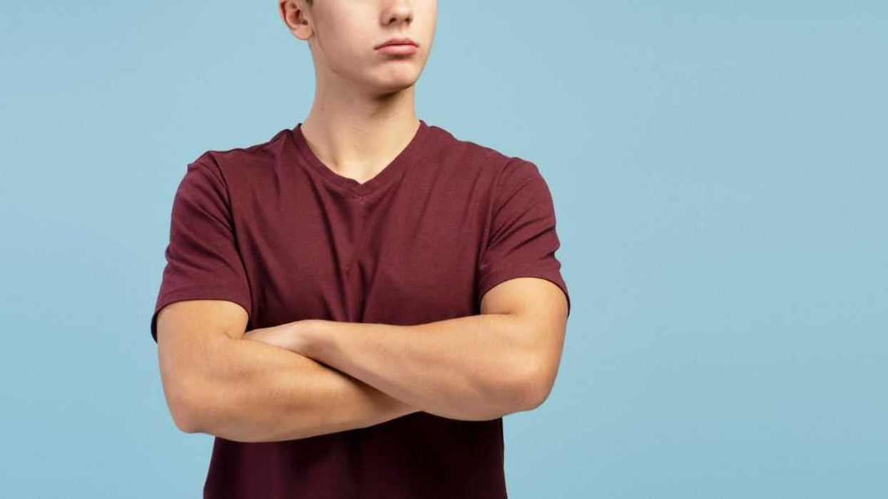 Serious, sad teen boy with crossed arms looking away on copy space, standing on blue background.