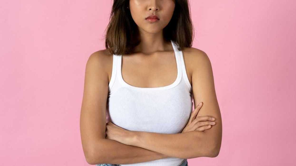 Serious young woman crossing arms and looking into camera against a plain pink background.