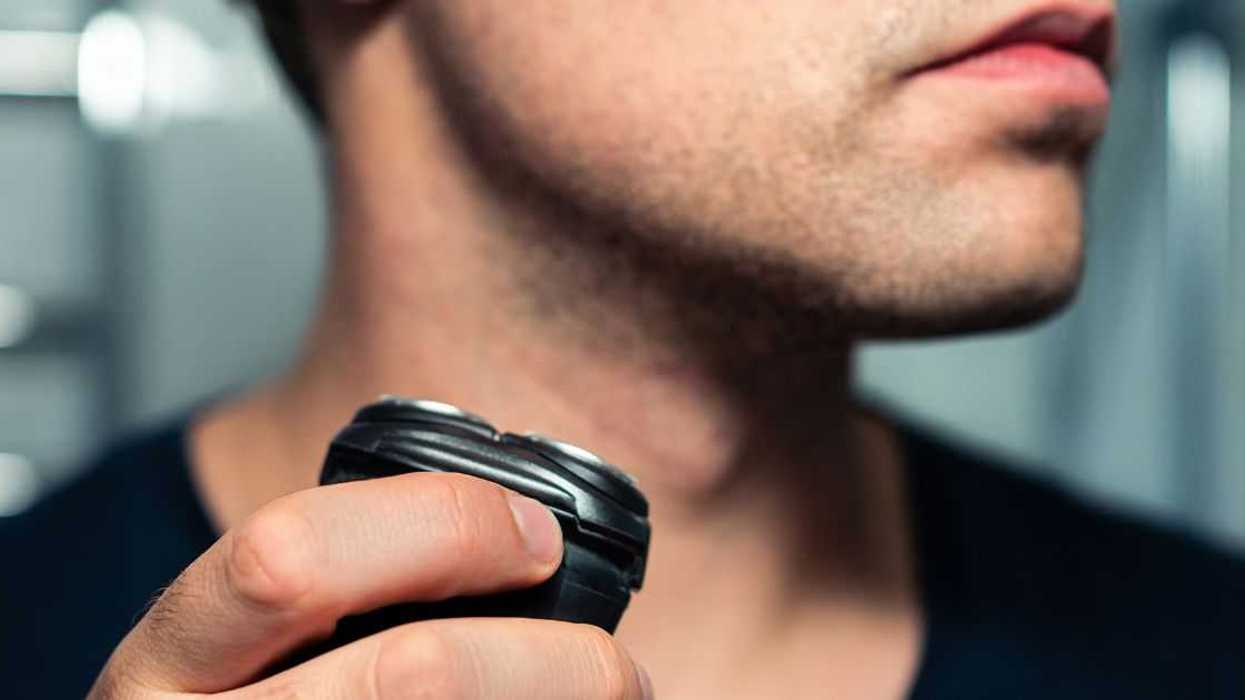 Shaving beard with machine. Man with electric shaver. Trimmer for face hair. Male grooming razor. Mirror in bathroom. Short stubble. Closeup of neck and chin.