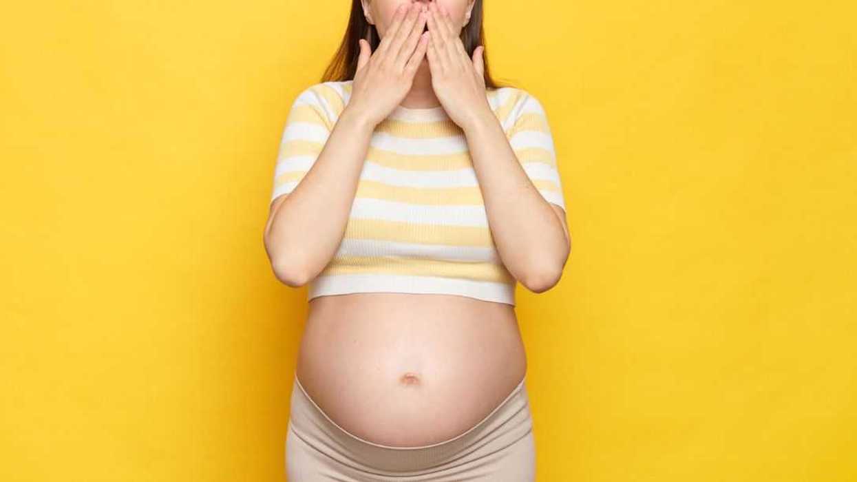 Shocked, astonished pregnant woman with bare belly wearing casual top isolated over yellow background screaming with amazement covering mouth with hands.