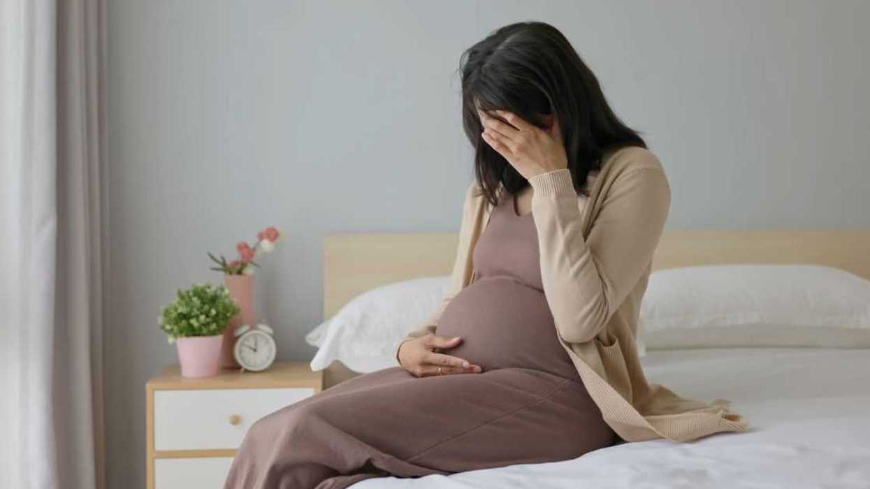 Side view of a pregnant woman touching her belly and sitting on a bed - she is feeling depressed and sad, crying in a bedroom.