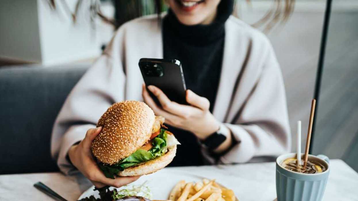 Smiling young woman sitting by the window in a restaurant enjoying her lunch. She is taking photos of her delicious food, fresh burger with fries with smartphone. Eating out lifestyle.
