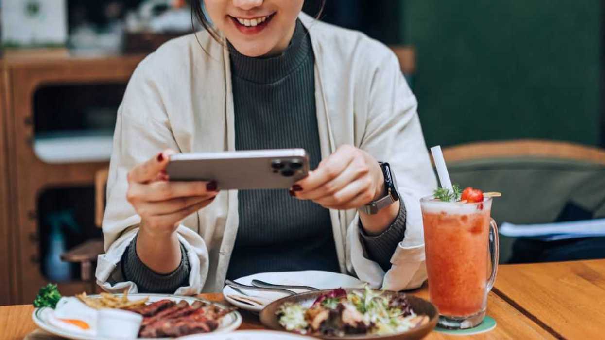 Smiling young woman taking photos of her scrumptious and beautifully plated dishes on the table with her smartphone in outdoor cafe. Enjoying her meal and sharing foodie photos on social media. Digital dining. People, food, lifestyle and technology.