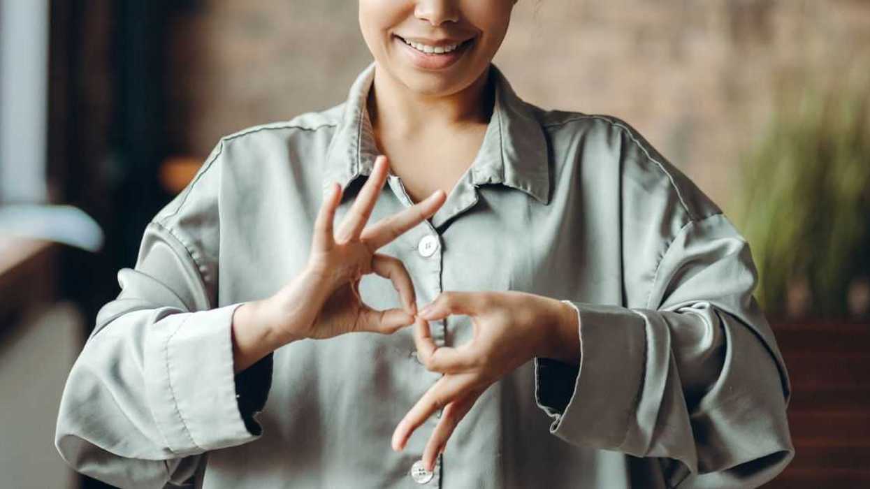 Smiling young woman using sign language, demonstrating the letter r with her hands while standing in a stylish modern loft apartment filled with natural light.
