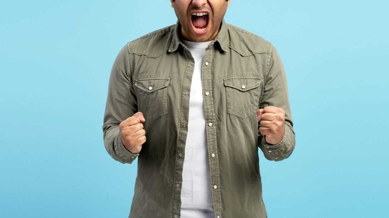 Studio portrait of an angry man clenching his fists and screaming with his eyes closed, expressing frustration and rage against a vibrant blue backdrop.