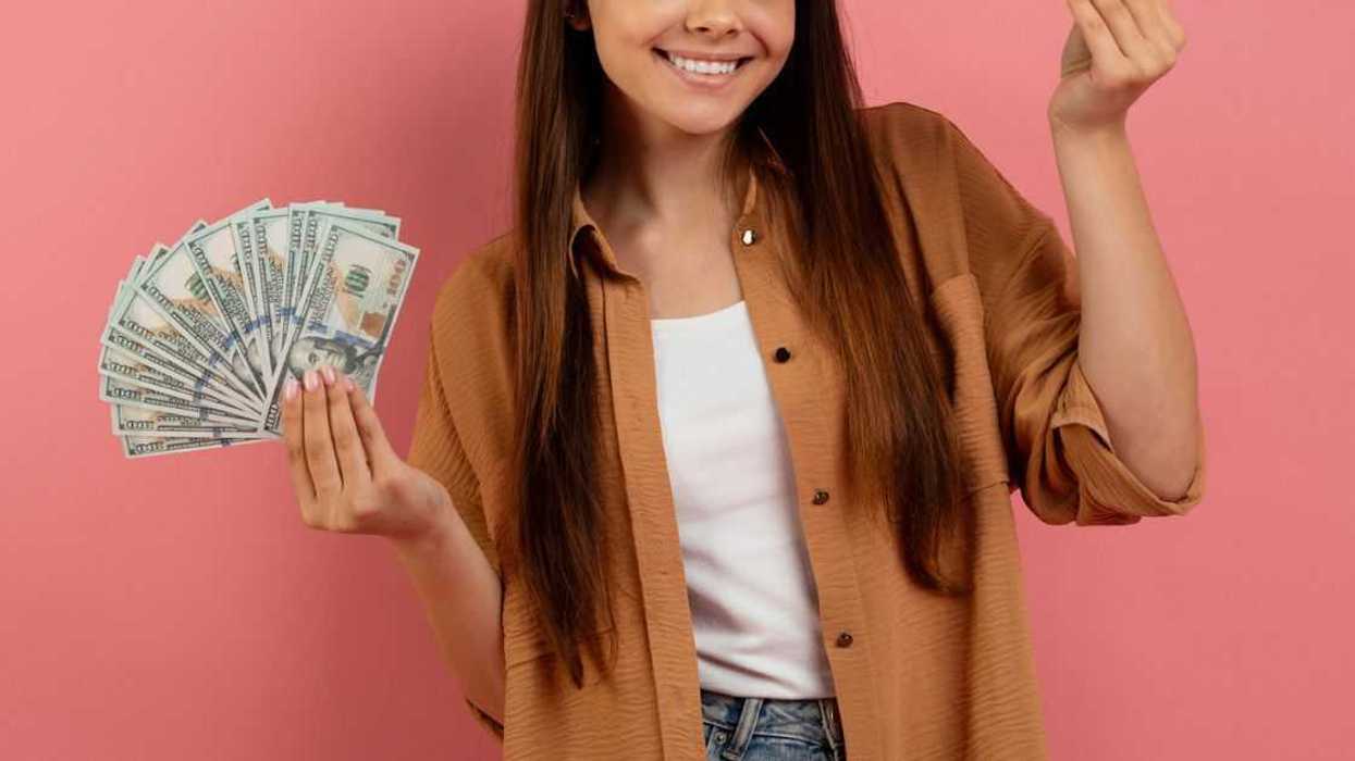 Teen Girl Holding Cash In Hands And Smiling At Camera. Standing In Front of a Pink Background.