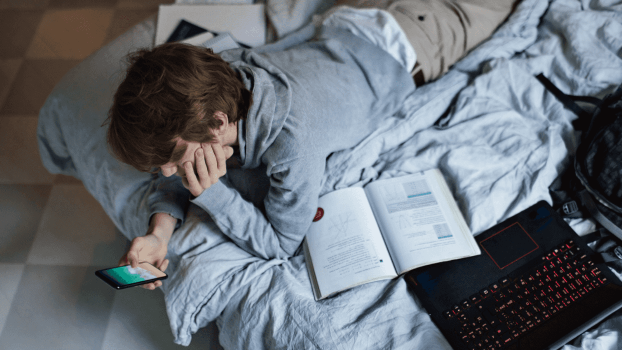 teen laying on bed with laptop and textbooks looking at phone